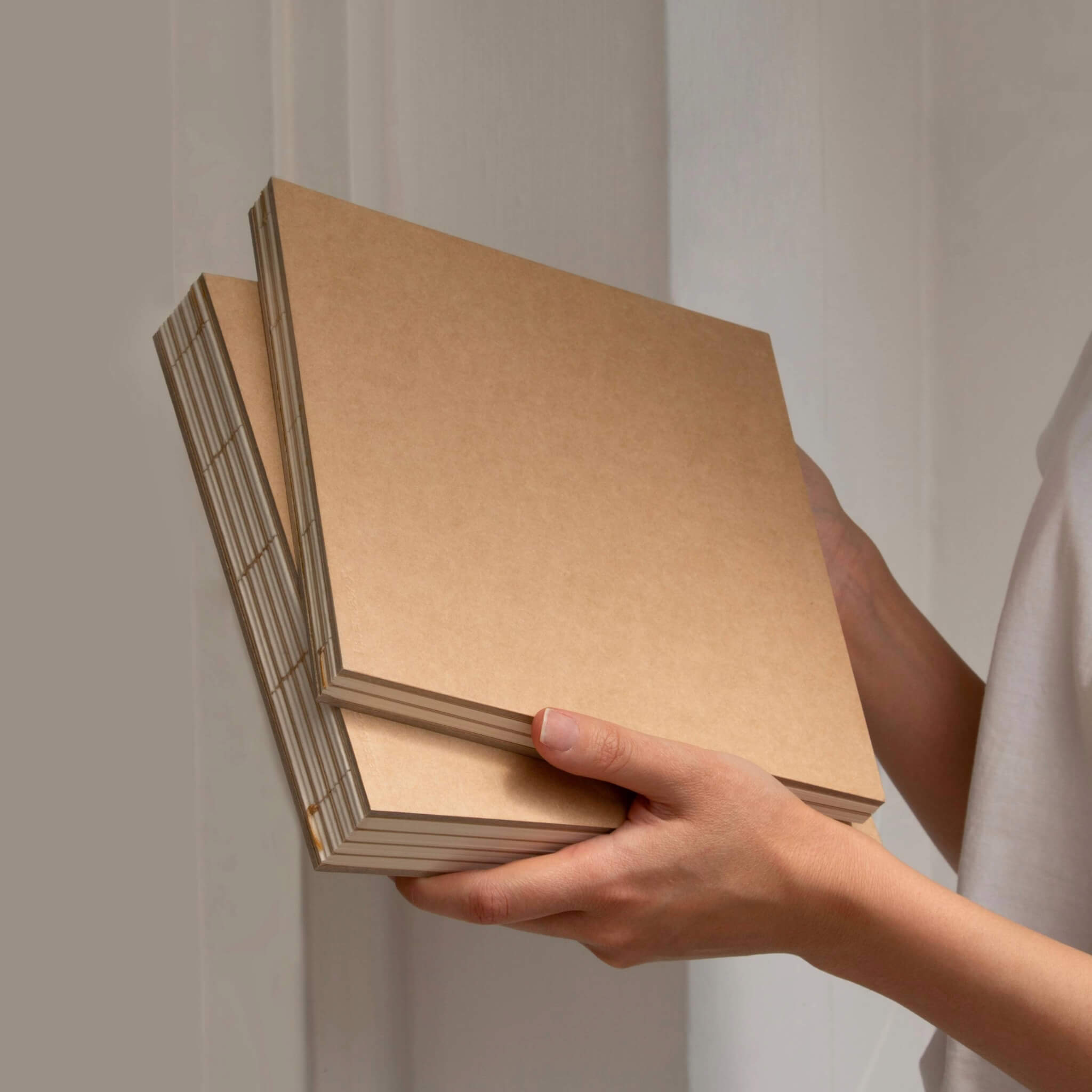 Hand-sewn 22 × 22 cm square sketchbook in natural kraft paper, shown in hand with exposed binding and layered pages