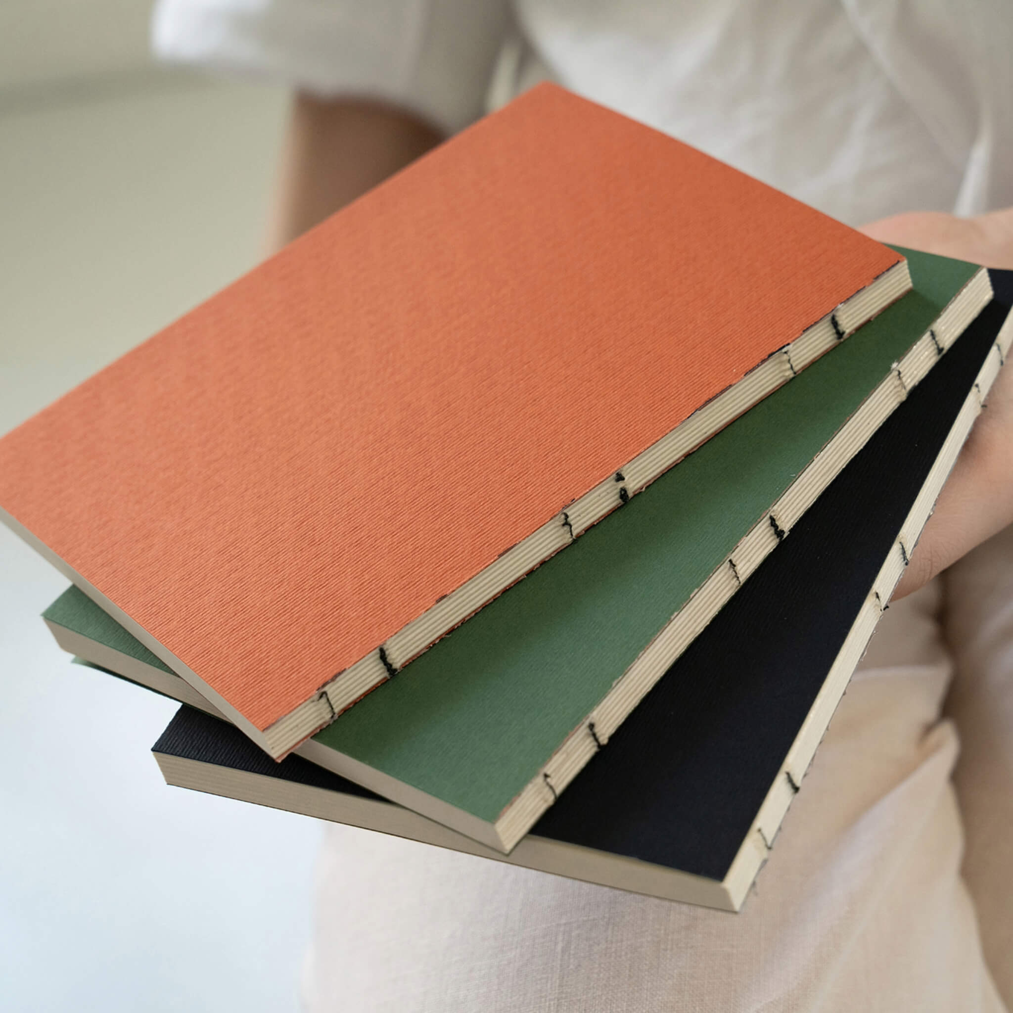 Stack of three basenotes 0144 Notebooks in orange, green, and black, showing exposed hand-sewn spines.