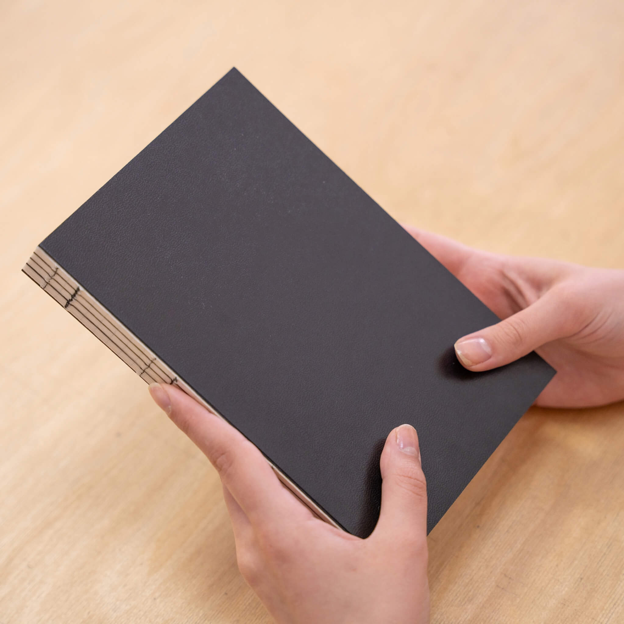 Hands holding the Basenotes 0432 A5 Notebook with a black cover, showcasing its exposed thread binding and minimalist design.