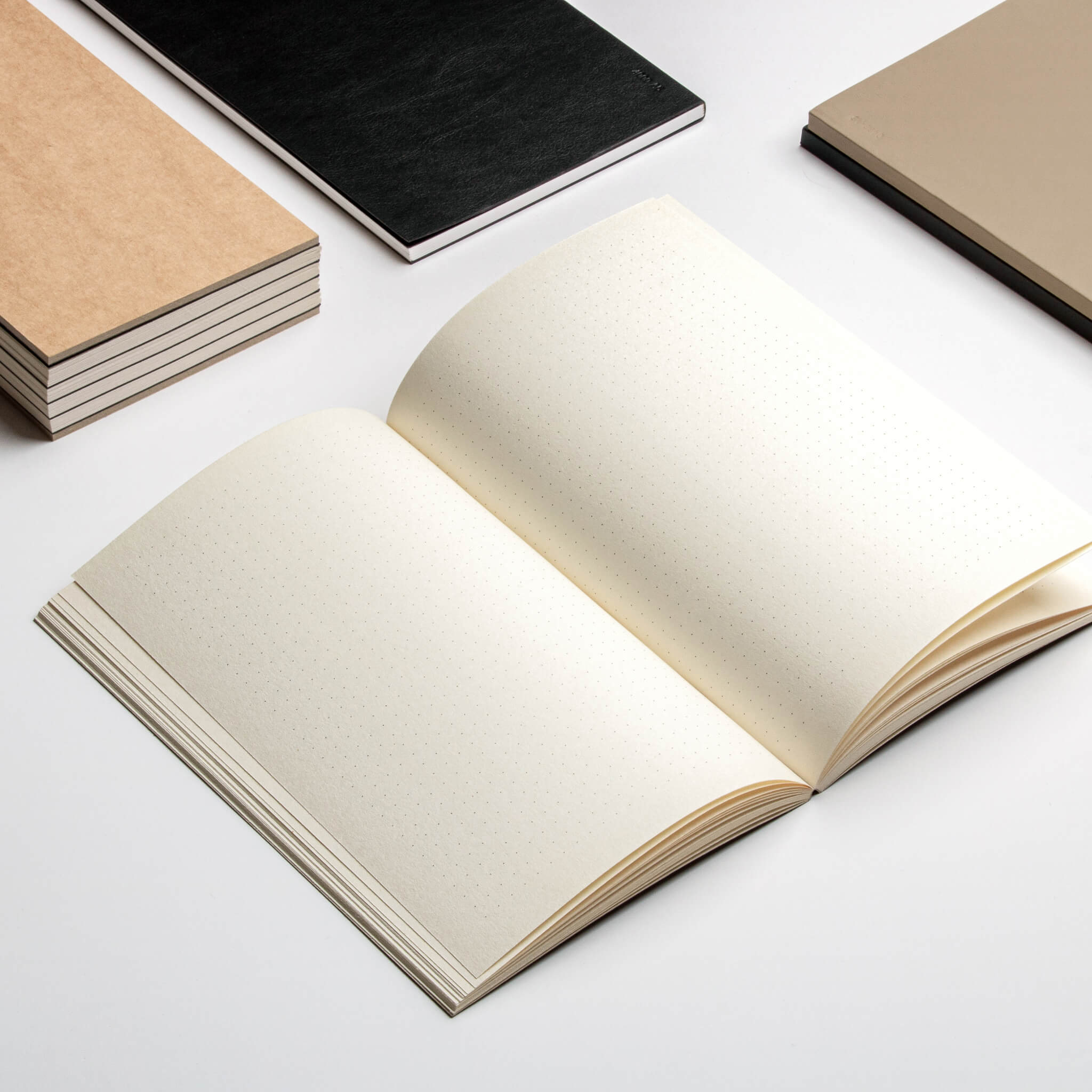 Open basenotes 0480-A5 Notebook showing dotted cream pages, placed beside black and beige notebooks on white surface.