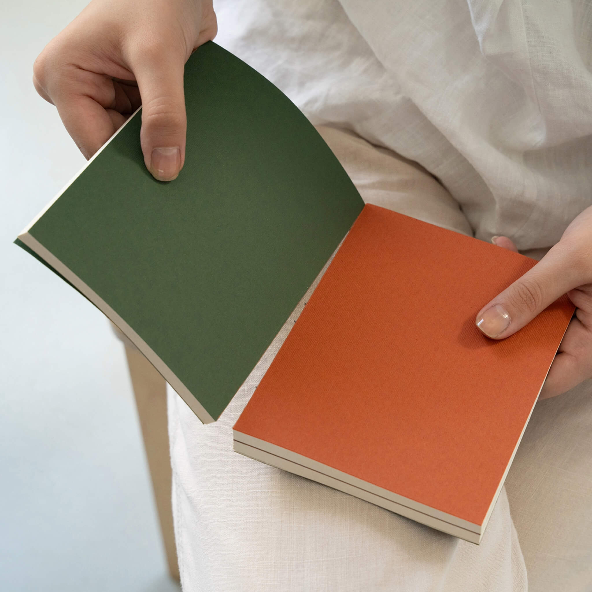 Person holding and opening basenotes 0144 Notebook showing contrasting green and orange covers with lined interior pages.