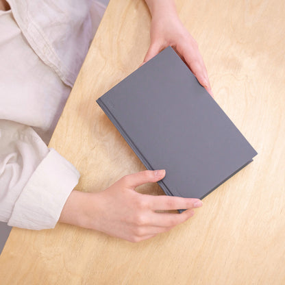 Hands holding a grey basenotes 0256 Notebook on a wooden surface, showing its clean cover and rounded spine.