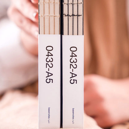 Close-up of two basenotes 0432 A5 Notebooks in kraft and black editions, showing packaging labels and exposed hand-stitched spines.