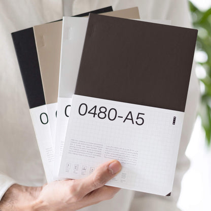 Person holding a stack of basenotes 0480-A5 Notebooks in beige, black, and grey covers, wrapped in the signature label sleeve.