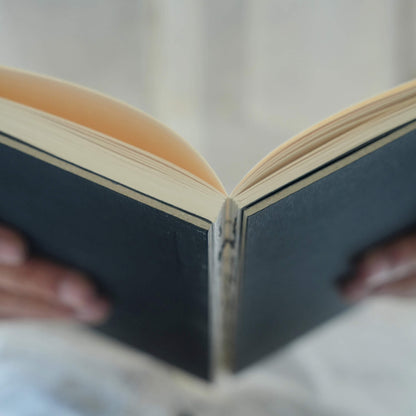 Hands holding an open basenotes 0880 A5 Notebook with a black cover, showcasing lay-flat exposed binding and cream pages.