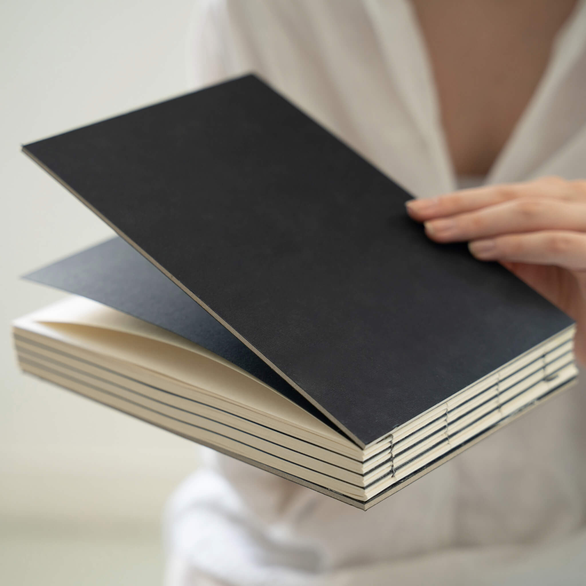 Person holding a stack of black basenotes 2316 sketchbooks showing exposed hand-stitched spines and cream-toned pages.