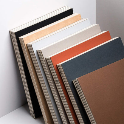 Line-up of basenotes 2333 Paperbooks in multiple colours black, kraft, rust, charcoal, mist, beige, and brown — all with exposed binding.