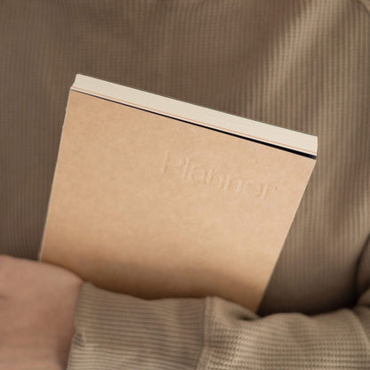 basenotes Planner in kraft cover with embossed title, held gently a minimalist design made with sustainable materials.